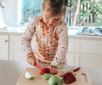 Niña utilizando el cuchillo de autonomía infantil KiddiKutter rosa para cortar fruta con supervisión adulta.
