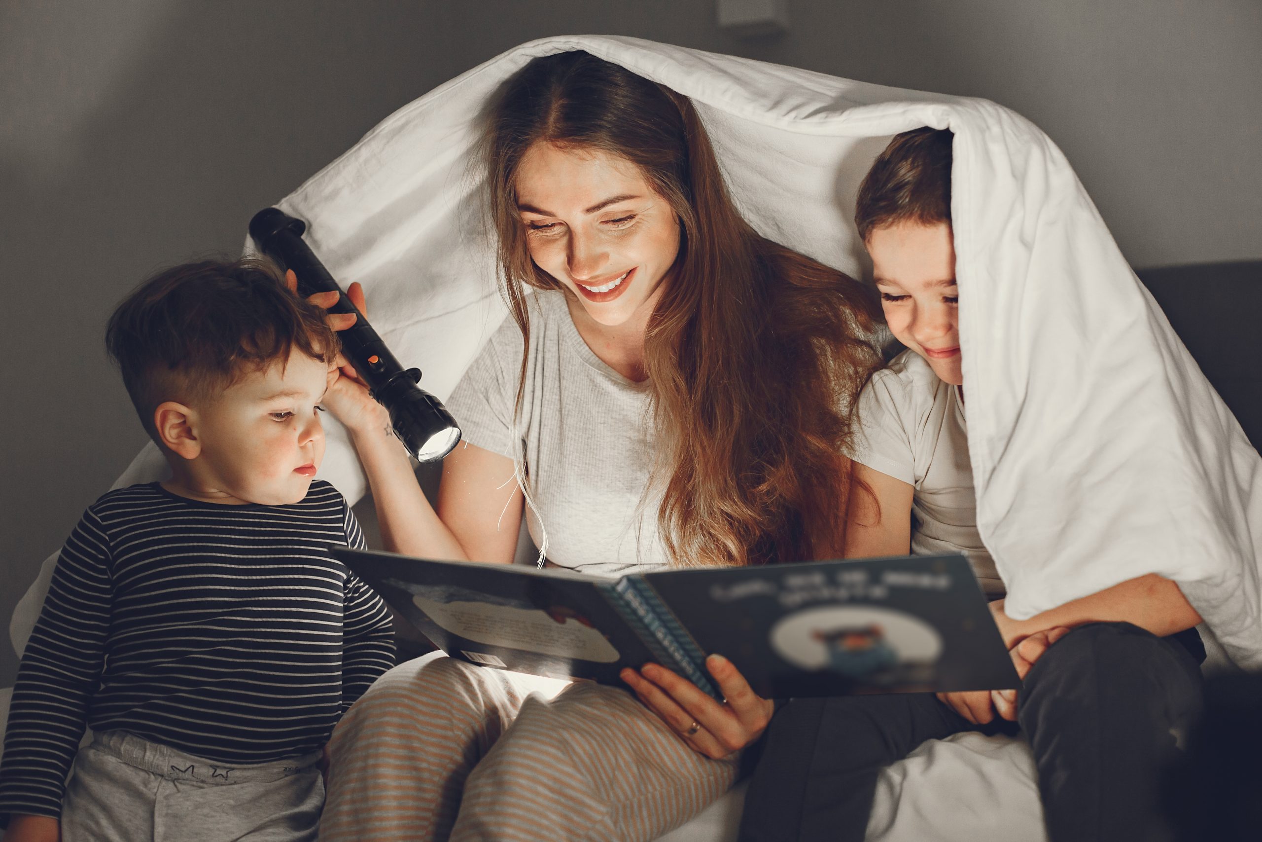 madre leyendo cuento a sus hijos en la cama bajo las sábanas momento de lectura en familia